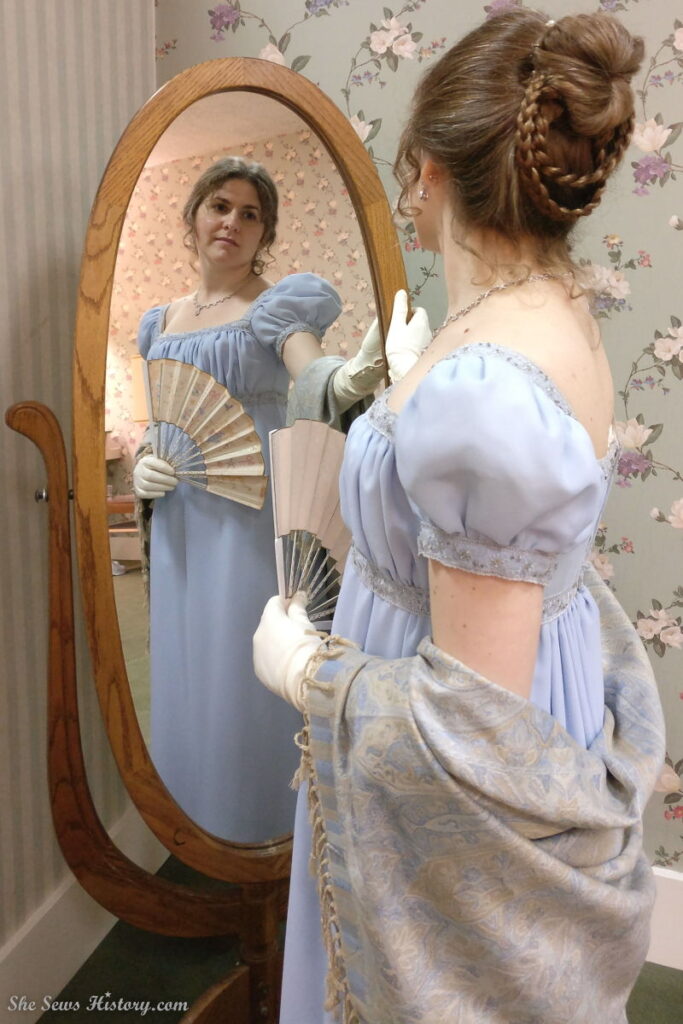 Girl in blue regency gown admires herself and hairstyle in mirror